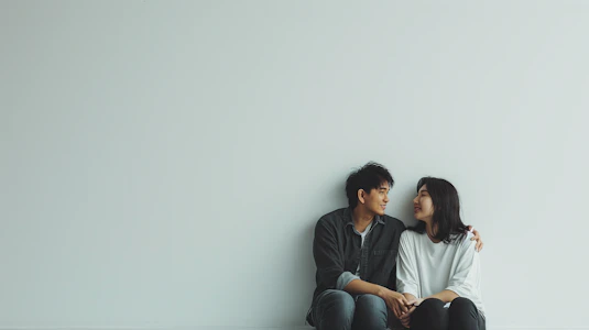 Young Asian couple sitting on the floor, smiling at each other against a minimalist white background. Photo by Pablo Stanley