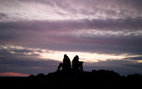 Silhouetted figures sit back-to-back against a dramatic purple and pink sky, creating a serene and introspective scene. Photo by Mikiwa